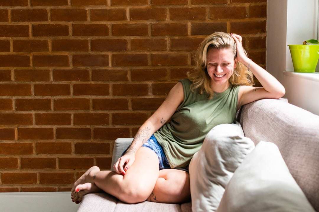 Uma mulher sentada em um sofá, sorrindo, com um objeto verde no fundo, luz natural suave.