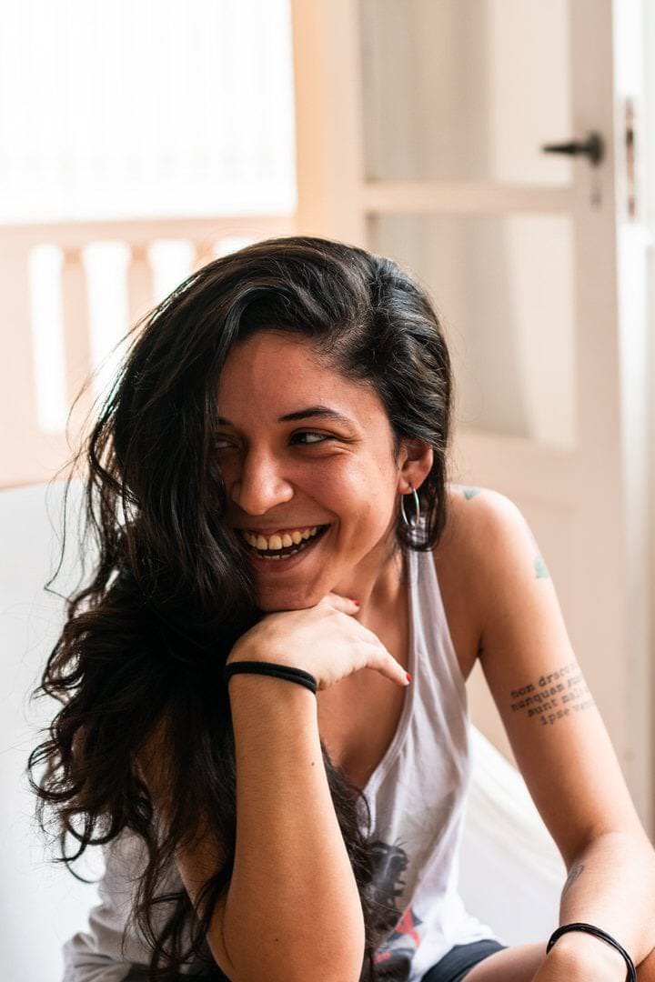 Uma mulher sentada frente a uma parede com porta no fundo, luz natural suave no canto inferior.