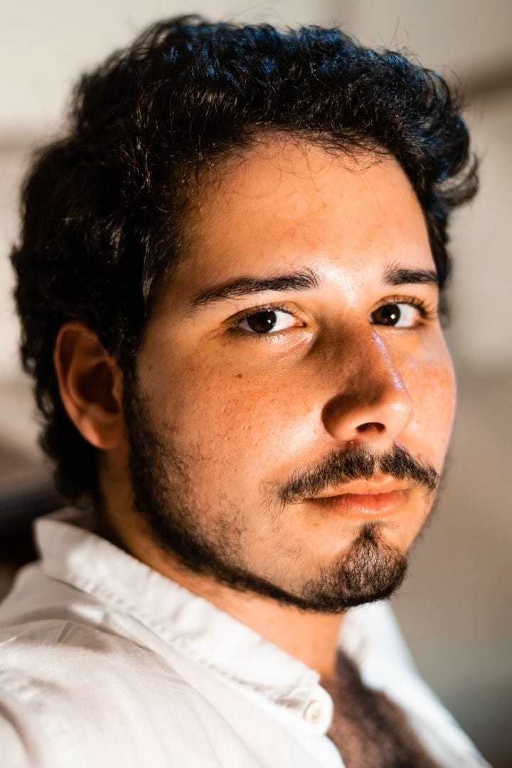 Um homem sentado em um banco de concreto, sob luz natural difusa, com a silhueta marcada por um céu claro.