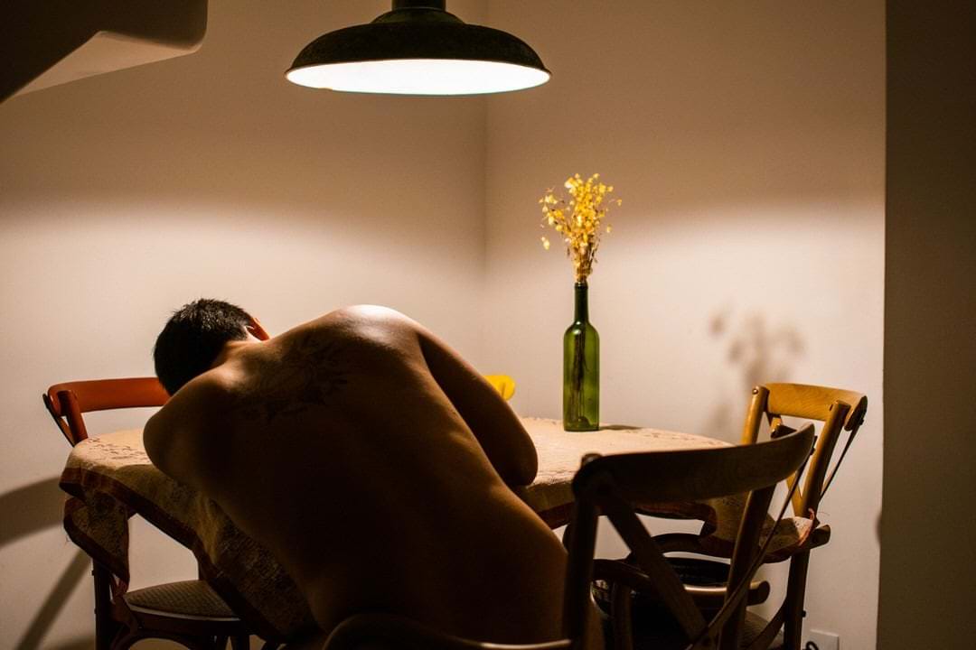 Um homem sentado em uma cadeira, frente a uma mesa com vaso de flor. Luz direta no teto.