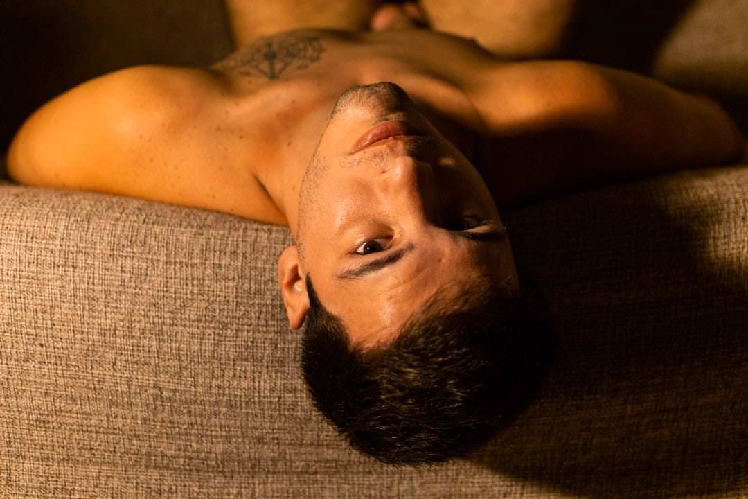 Pessoa de costas sentada em sofá, olhos voltados para a câmera, luz natural suave vindo da janela lateral.