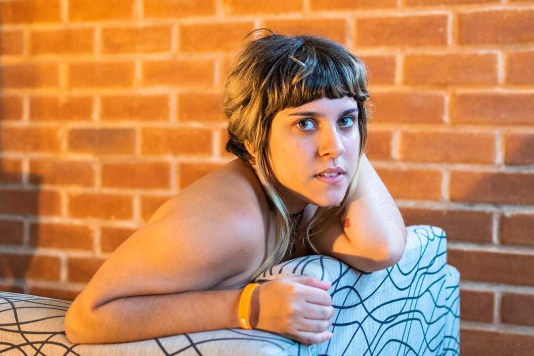 Uma mulher sentada em um sofá, olhando para a parede de fundo, iluminada por luz natural suave.