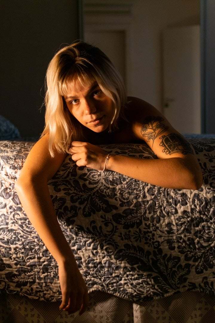 Uma mulher sentada em cama, veste vestido preto, com fundo de parede.