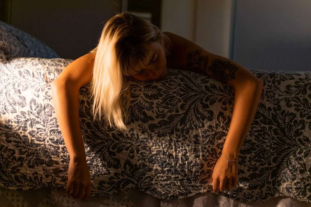 Uma mulher debruçada em cama, veste vestido preto e branco, luz natural sussurra por cima da cabeceira.