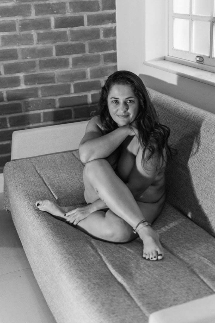 Uma mulher sentada em um sofá, com um fundo de parede de tijolo e uma janela.
