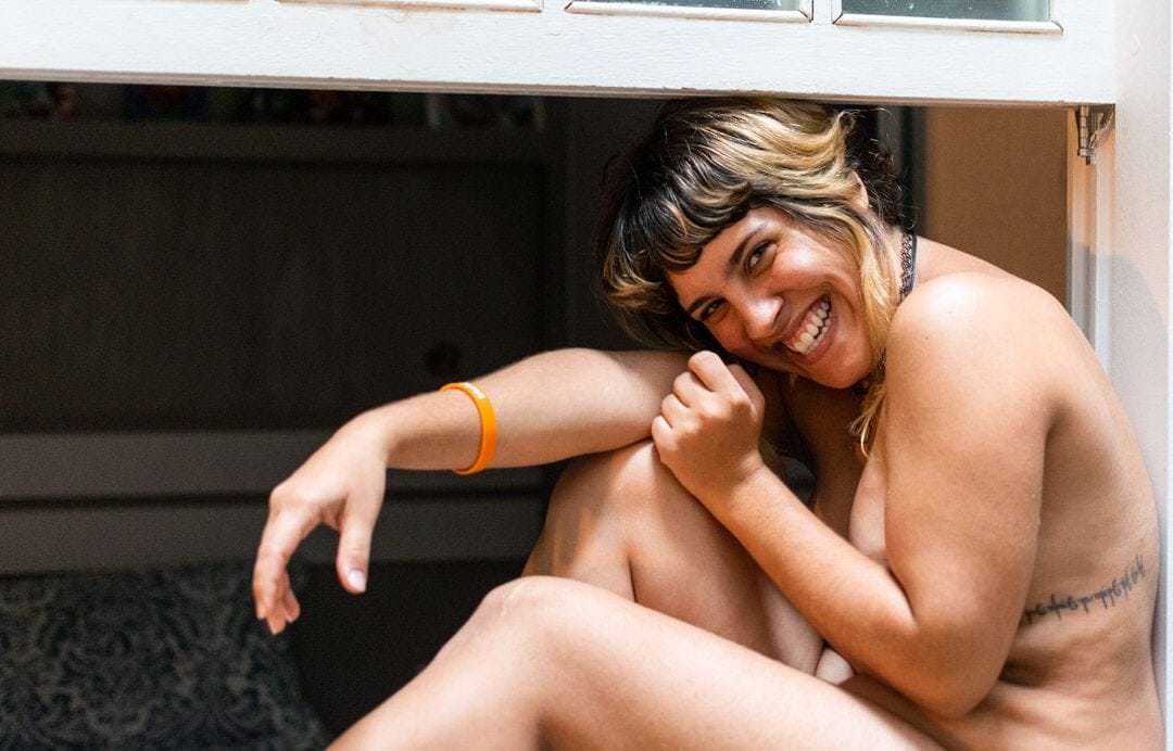 Uma mulher sentada e sorrindo diante de um fundo com parede reta e luminoso.