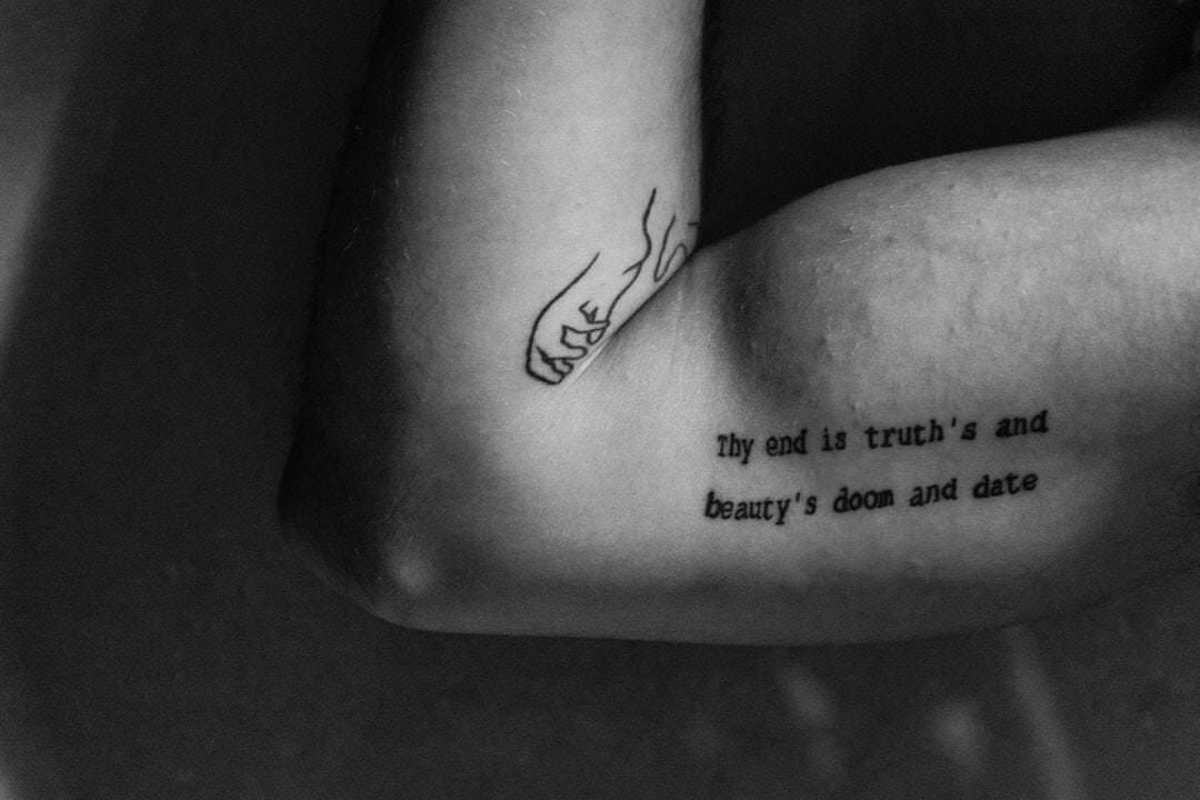 Mãos com tatuagem visível em ambiente escuro, luz difusa de fundo, detalhe em contraste com fundo suave.