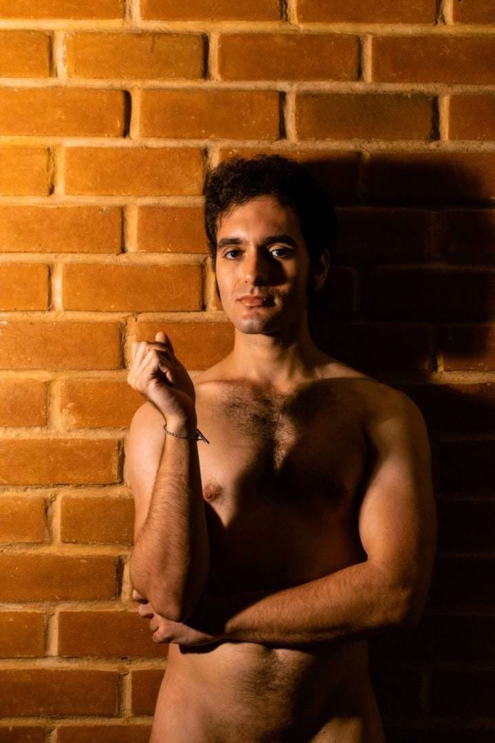 Pessoa em pé diante de um muro, luz natural difusa no fundo.