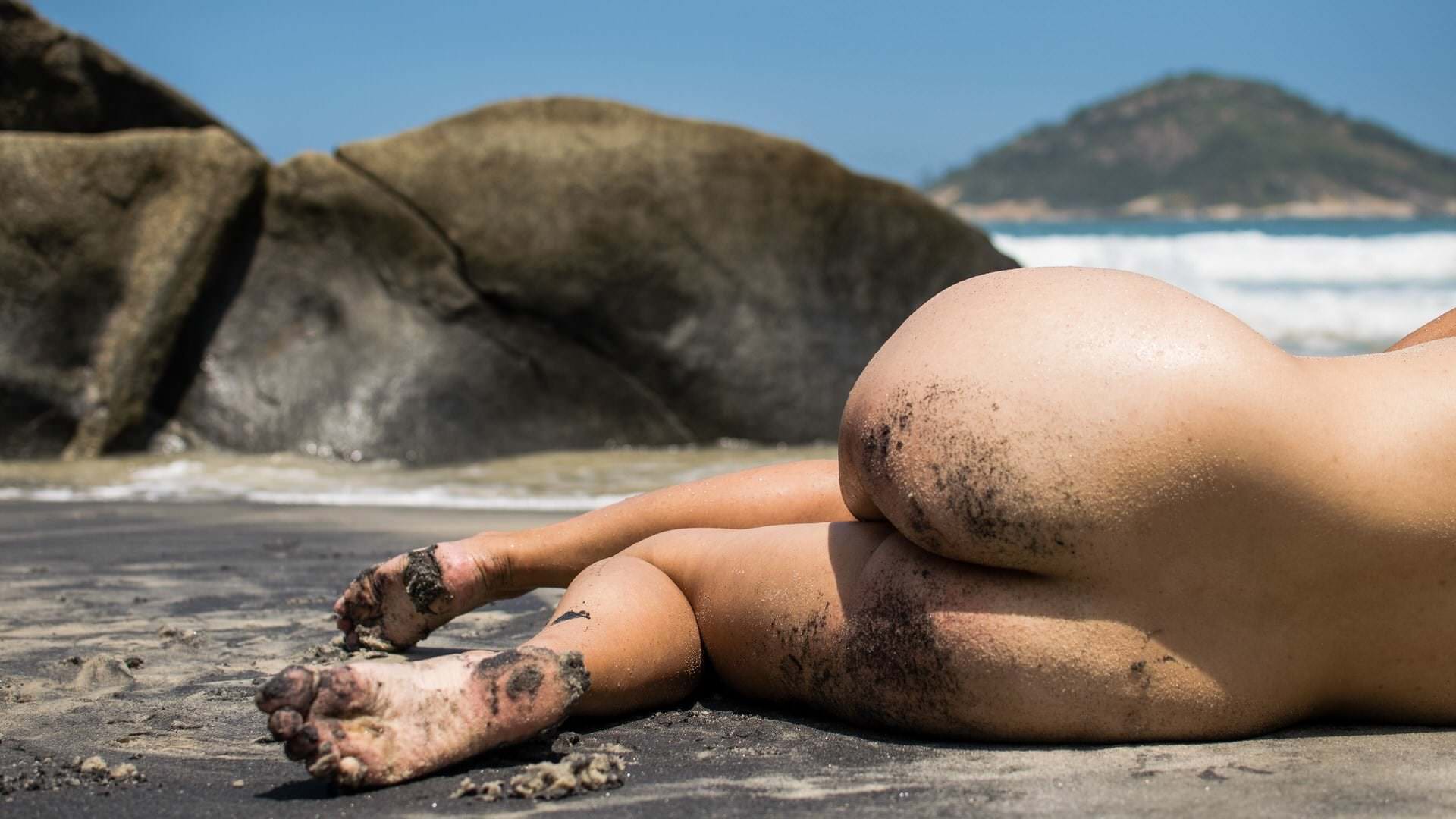 Pessoa de costas sobre o chão, cercada por rochas, água, colinas e céu aberto, iluminação natural suave.