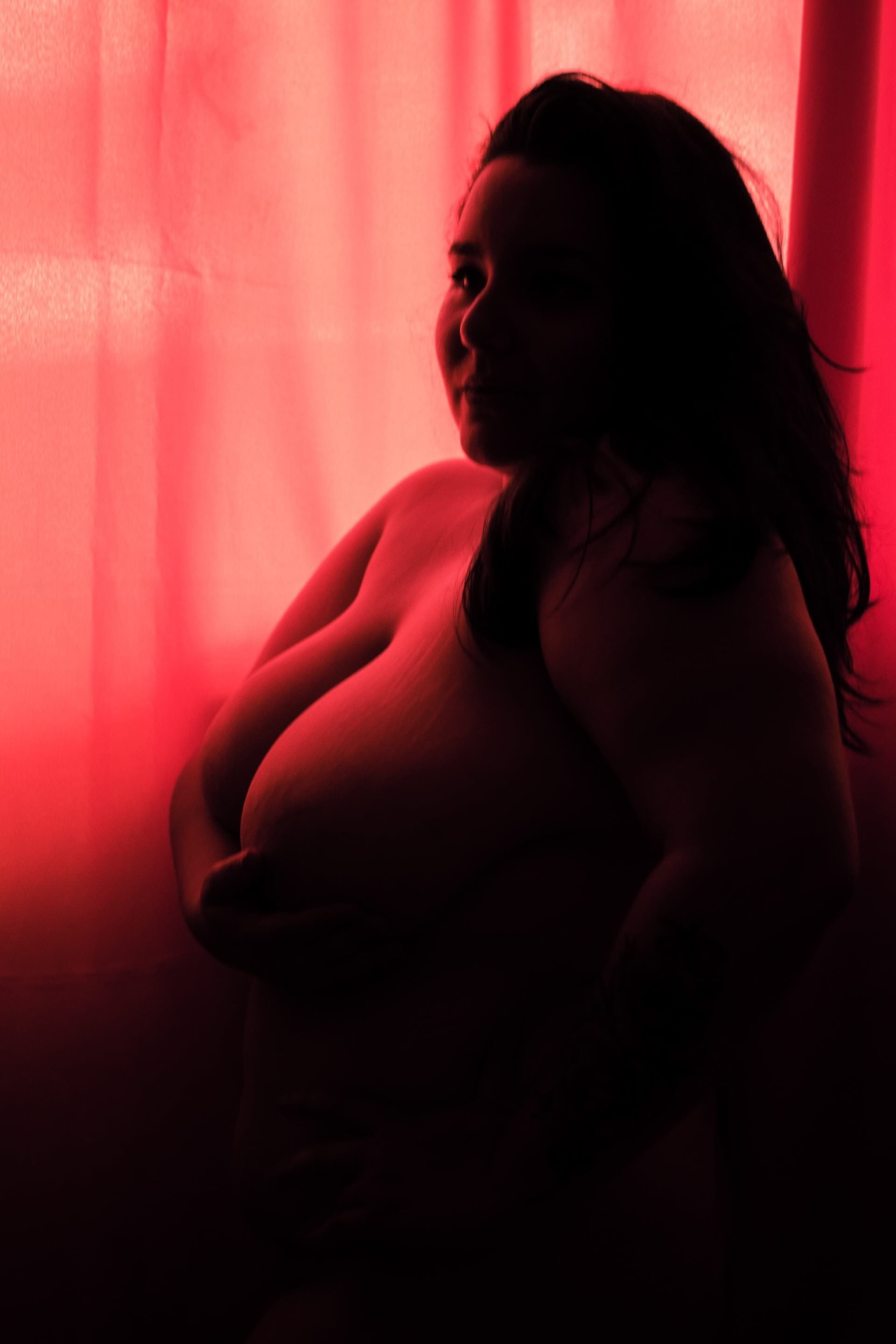 Uma mulher sentada, olhando para a câmera, com um tecido de cortina atrás de si, iluminada por luz natural suave.