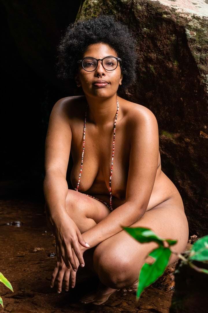 Mulher com óculos sentada sobre uma rocha, fundo com solo abaixo.