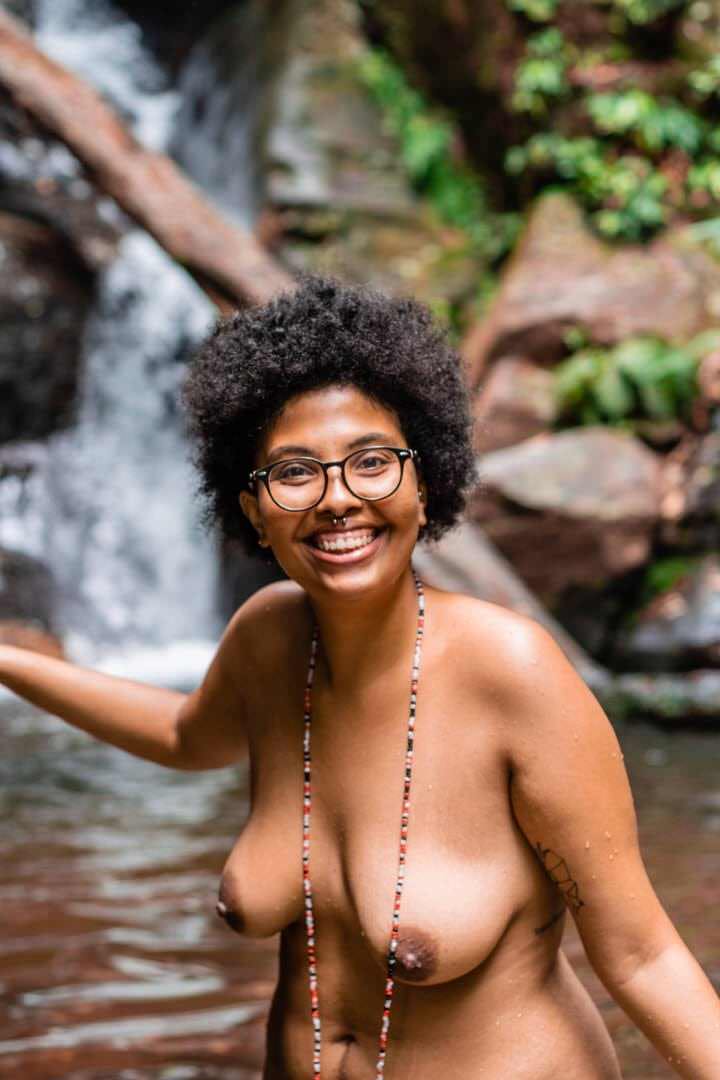 Uma mulher de costas, em frente a uma queda d'água, com rochas visíveis no fundo, luz natural suave e contraste entre a água e a pedra.