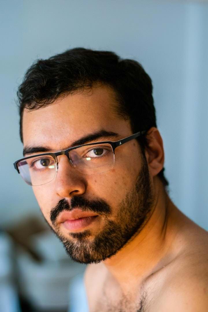 Homem com óculos de ação, sentado em banco de madeira, sob luz natural fraca e difusa.