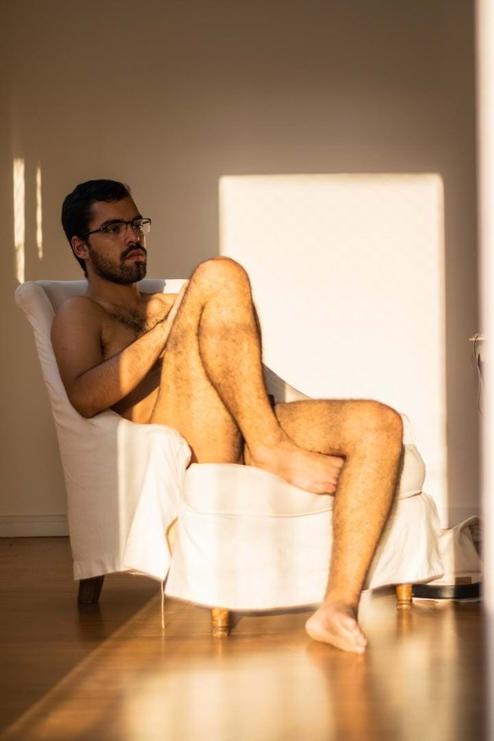 Pessoa sentada em cadeira, usando óculos, fundo com parede retangular, luz natural difusa.
