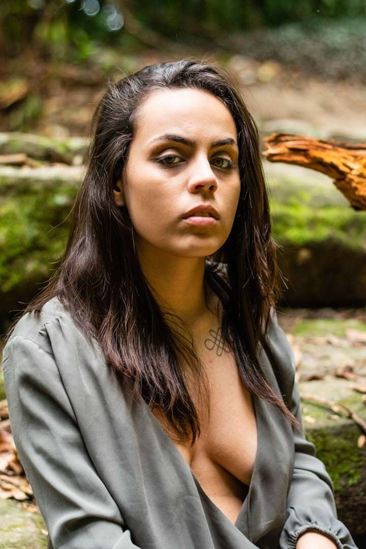 Uma mulher veste um vestido em tons de cinza, frente a árvores verdes no fundo, luz natural suave e composição equilibrada.