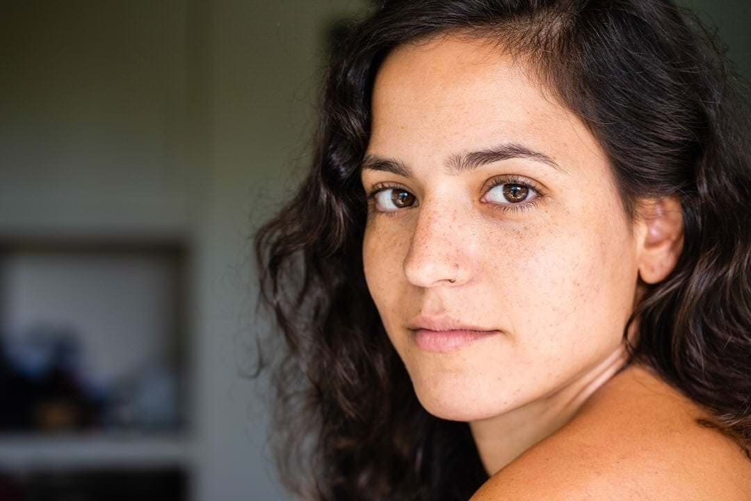 Uma mulher sentada em um banco, olhando para a frente, com fundo desfocado.