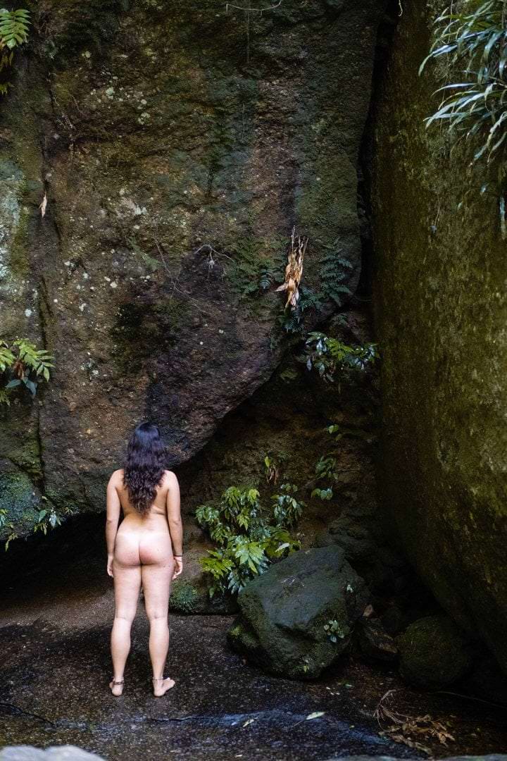 Uma mulher de costas, em pé sobre o solo, com rocha e vegetação no fundo.