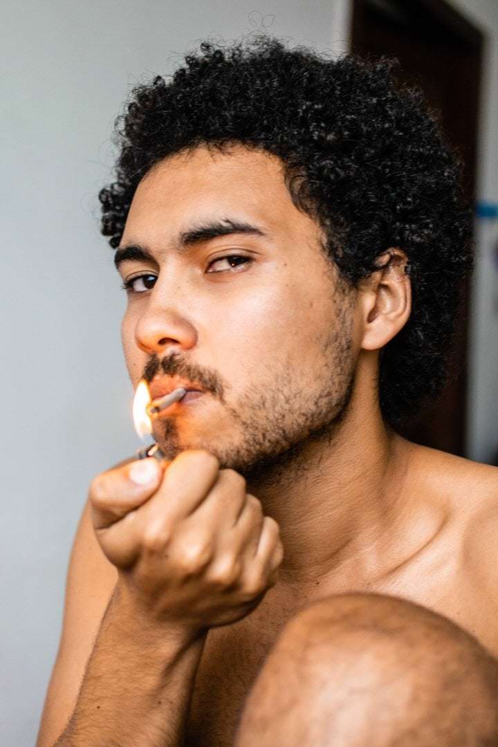 Um homem segura um cigarro em sua mão, em um ambiente escuro com luz difusa vinda de uma janela.
