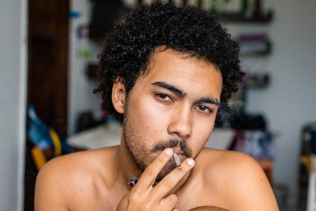 Um homem segura um cigarro enquanto um muro de fundo está iluminado por luz natural fraca.