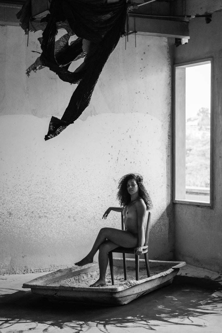 Uma mulher sentada em uma cadeira, ambiente em preto e branco, luz difusa e contraste marcado.