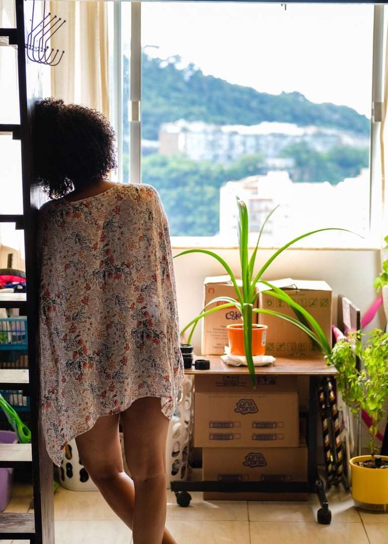 Uma mulher está em frente a caixas e plantas verdes, com árvores verdes no fundo e céu branco.