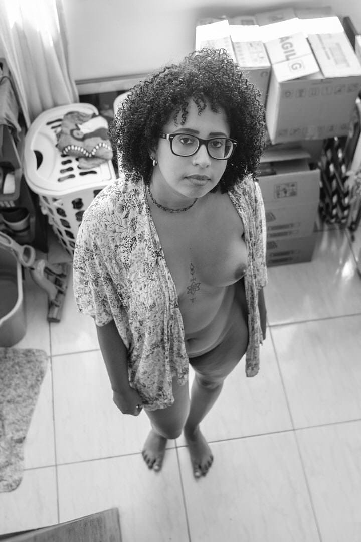 Uma mulher com óculos está em pé no chão, cercada por caixas, roupas e objetos no fundo.