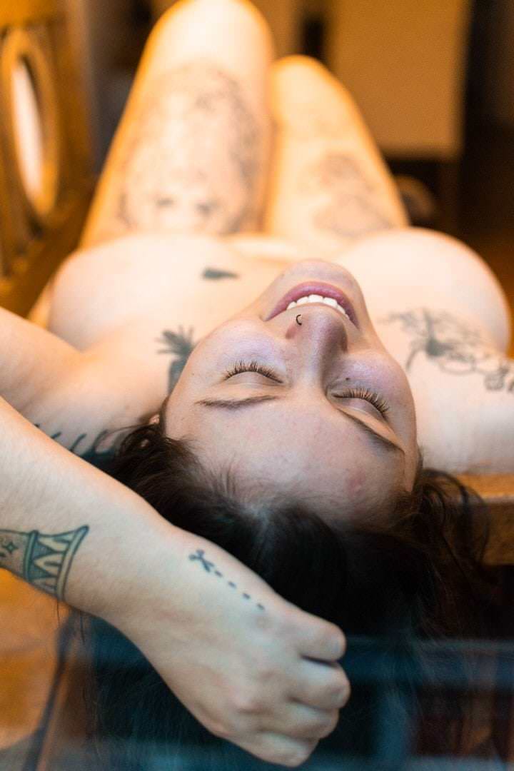 Uma mulher de costas está deitada em uma cama, com roupas soltas e luz natural saindo da janela.