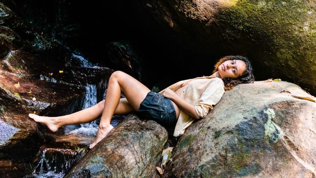 Uma mulher de costas repousa sobre uma pedra, com cascata de água no fundo, luz difusa e contraste natural.