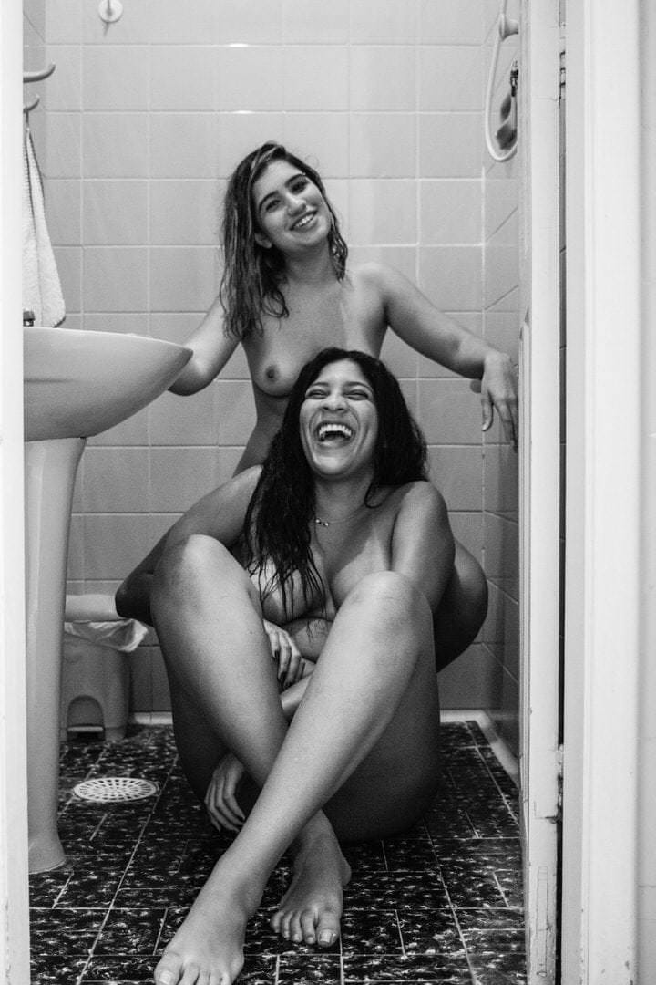 Duas mulheres sentadas no chão, próximo a um lavatório e uma toalha sobre a pia.