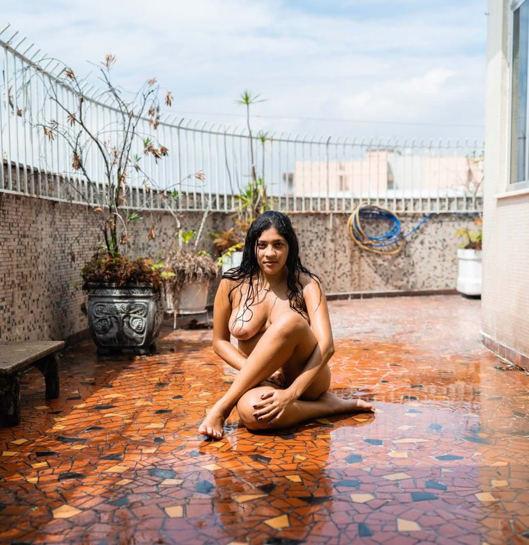 Uma mulher sentada no chão, cercada por plantas em vasos, um banco, uma grade e um tubo, com céu nublado no fundo.