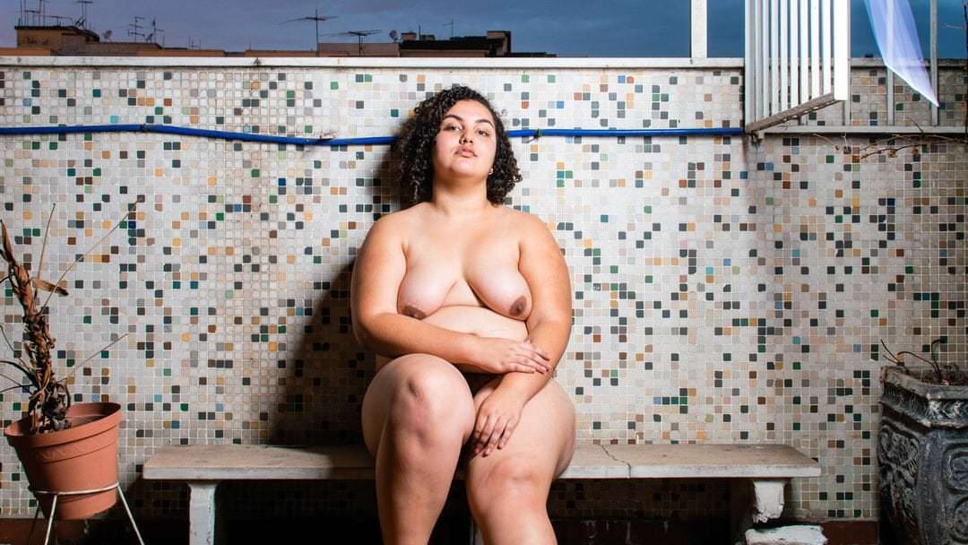 Uma mulher sentada em uma bancada, com um vaso e uma roupa sobre ele, em ambiente urbano com céu claro.
