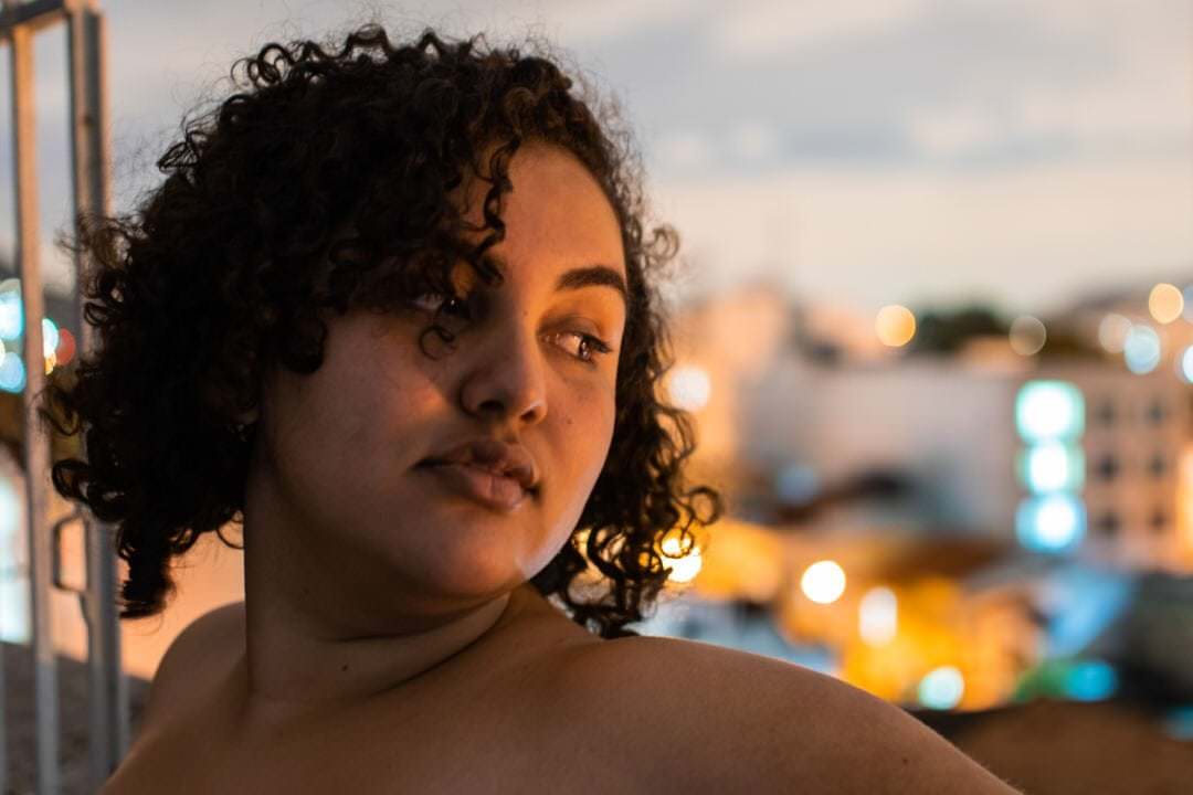 Uma mulher está a lado de um tubo metálico, com edifícios e luzes no fundo.