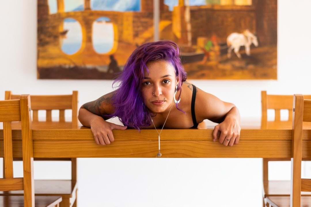 Uma mulher sentada em uma cadeira, com outras cadeiras ao redor e um quadro na parede de fundo.
