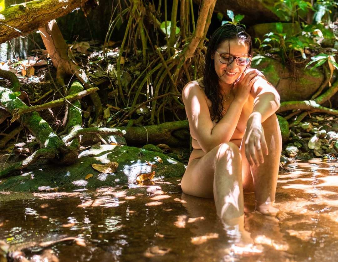 Uma mulher com óculos sentada sobre a superfície d'água, com árvores de fundo.