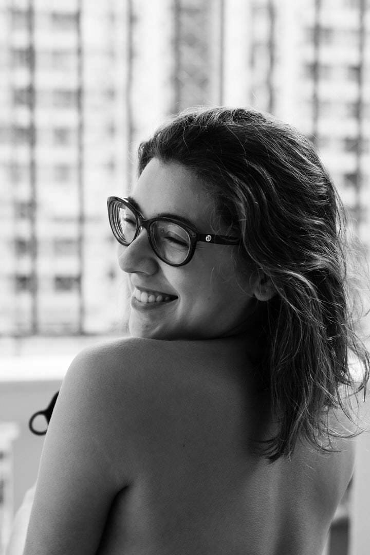 Uma mulher com óculos pretos está em frente a edifícios brancos, sob luz natural difusa.