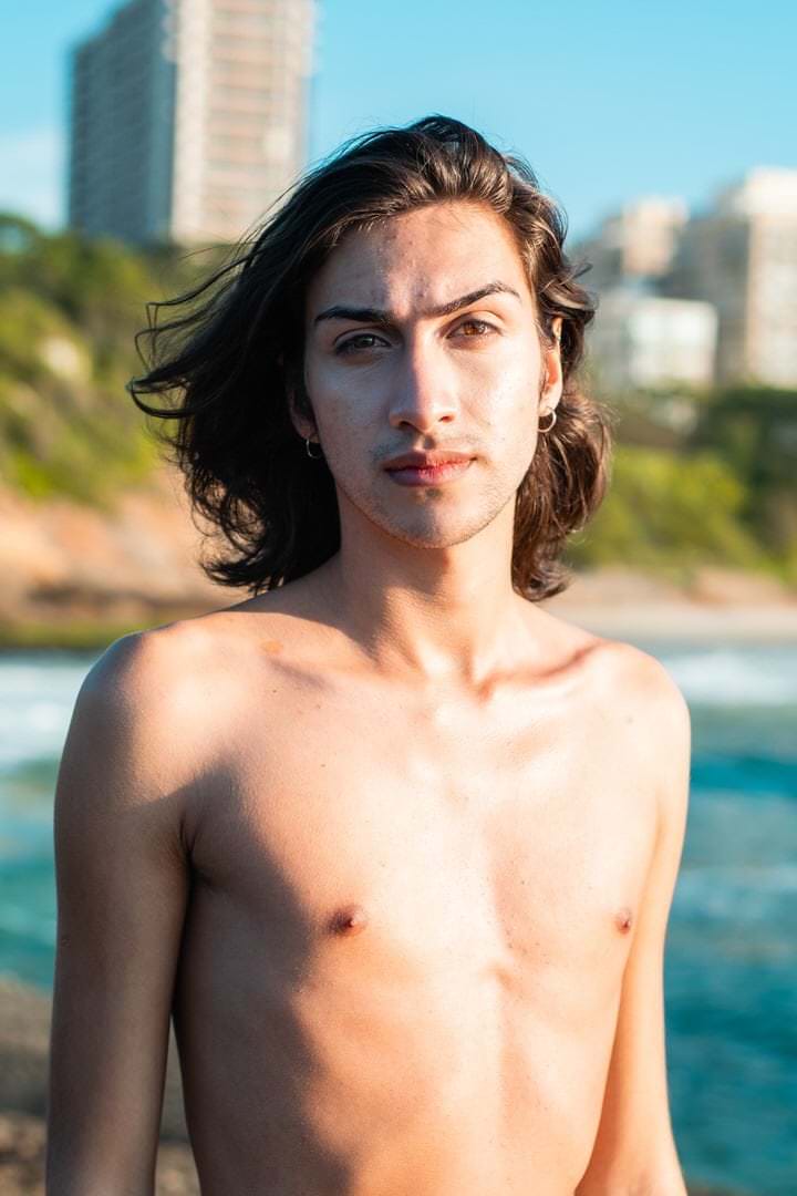 Um homem caminha em frente a edifícios, com céu claro no fundo, luz natural suave e ângulo amplo.