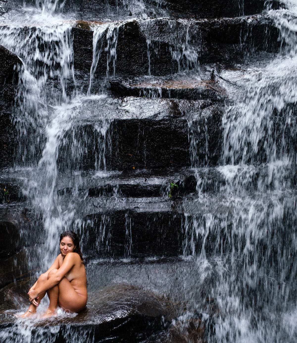 Uma mulher sentada em uma pedra, fundo com uma queda d'água em fundo claro e contrastado.