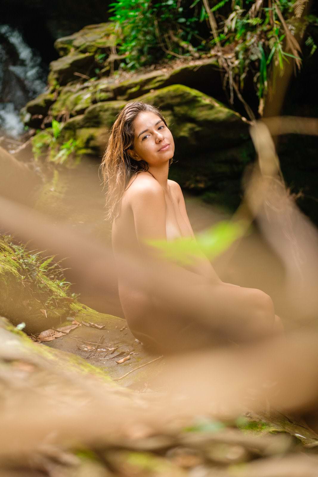 Uma mulher sentada em um banco, com árvores no fundo, luz natural suave e detalhes claros no ambiente.